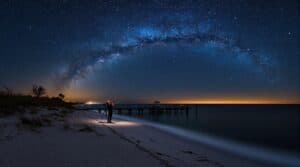 Person stargazing on a sandy Gulf Coast beach at night with telescope, sea oats in silhouette, Milky Way and stars bright in the sky, gentle waves reflecting starlight.