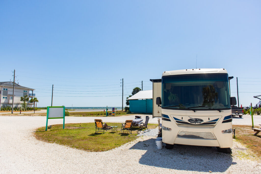 A white RV is parked on a gravel lot near the beach, with two empty camping chairs, a table, and power lines visible under a clear blue sky. A house and the ocean are in the background.