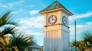 Classic white wooden clock tower with verdigris copper roof, framed by palm fronds under a clear blue sky, evoking small-town coastal charm.
