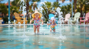 Two toddlers in swimsuits play at a seaside-themed splash pad with water jets, palm fronds, and colorful chairs in the background, under bright coastal sunlight.