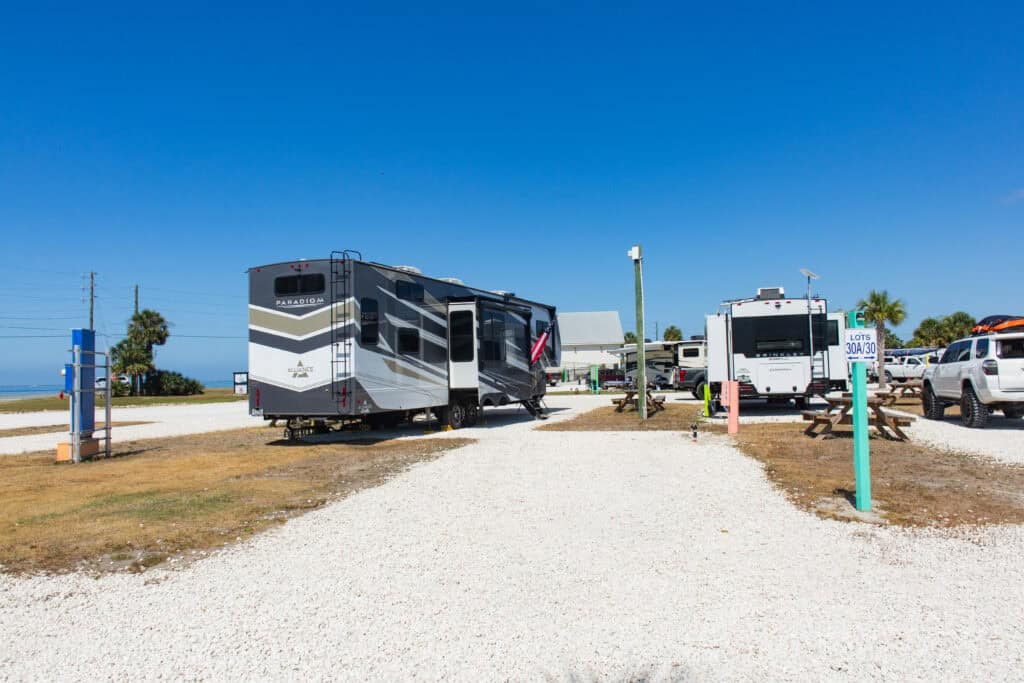 An RV park with gravel lots, several parked RVs, picnic tables, and a clear blue sky overhead.