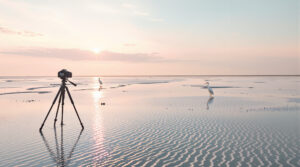 A DSLR camera on a tripod stands on a coastal salt flat at low tide, facing rippled sand and shallow pools reflecting a pastel sunrise sky, with a white egret in the distance and soft natural light illuminating the scene.