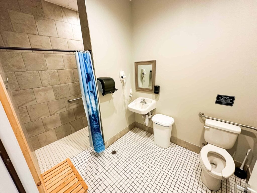 Accessible bathroom with a tiled shower, blue curtain, wooden bench, sink, mirror, paper towel dispenser, waste bin, and toilet with grab bars.