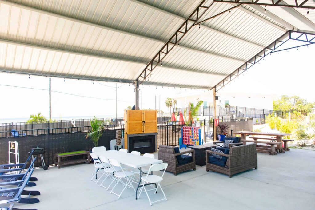 Covered outdoor patio with tables, chairs, sofas, and a fireplace. A large Connect Four game and exercise equipment are visible. String lights hang from the metal roof structure.