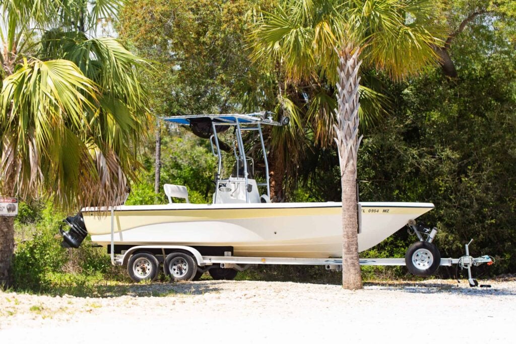 A small yellow and white motorboat on a trailer is parked on gravel beside palm trees and greenery.