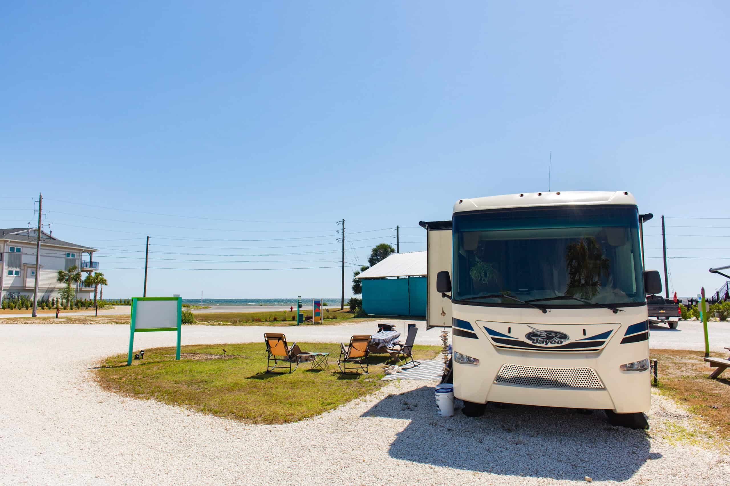 A white RV is parked at a campground near the beach with two empty chairs and a small table set up outside on a sunny day.