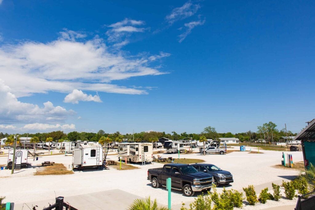 A sunny RV park with several parked trailers and trucks on gravel lots, surrounded by trees and under a partly cloudy blue sky.
