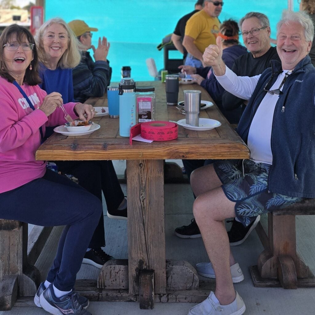 Five older adults sit at a wooden picnic table outdoors eating a meal, smiling, and giving a thumbs up. Bright sunlight and turquoise water are visible in the background.